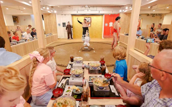 Family watching sumo show with retired wrestler at Ryogoku, enjoying chanko-nabe and mochi.