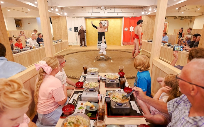Family watching sumo show with retired wrestler at Ryogoku, enjoying chanko-nabe and mochi.