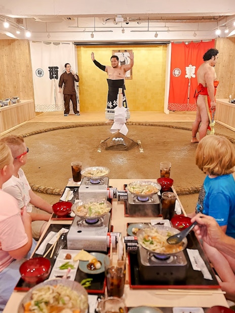 Family watching sumo show with retired wrestler at Ryogoku, enjoying chanko-nabe and mochi.