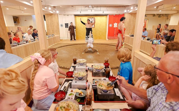 Family watching sumo show with retired wrestler at Ryogoku, enjoying chanko-nabe and mochi.