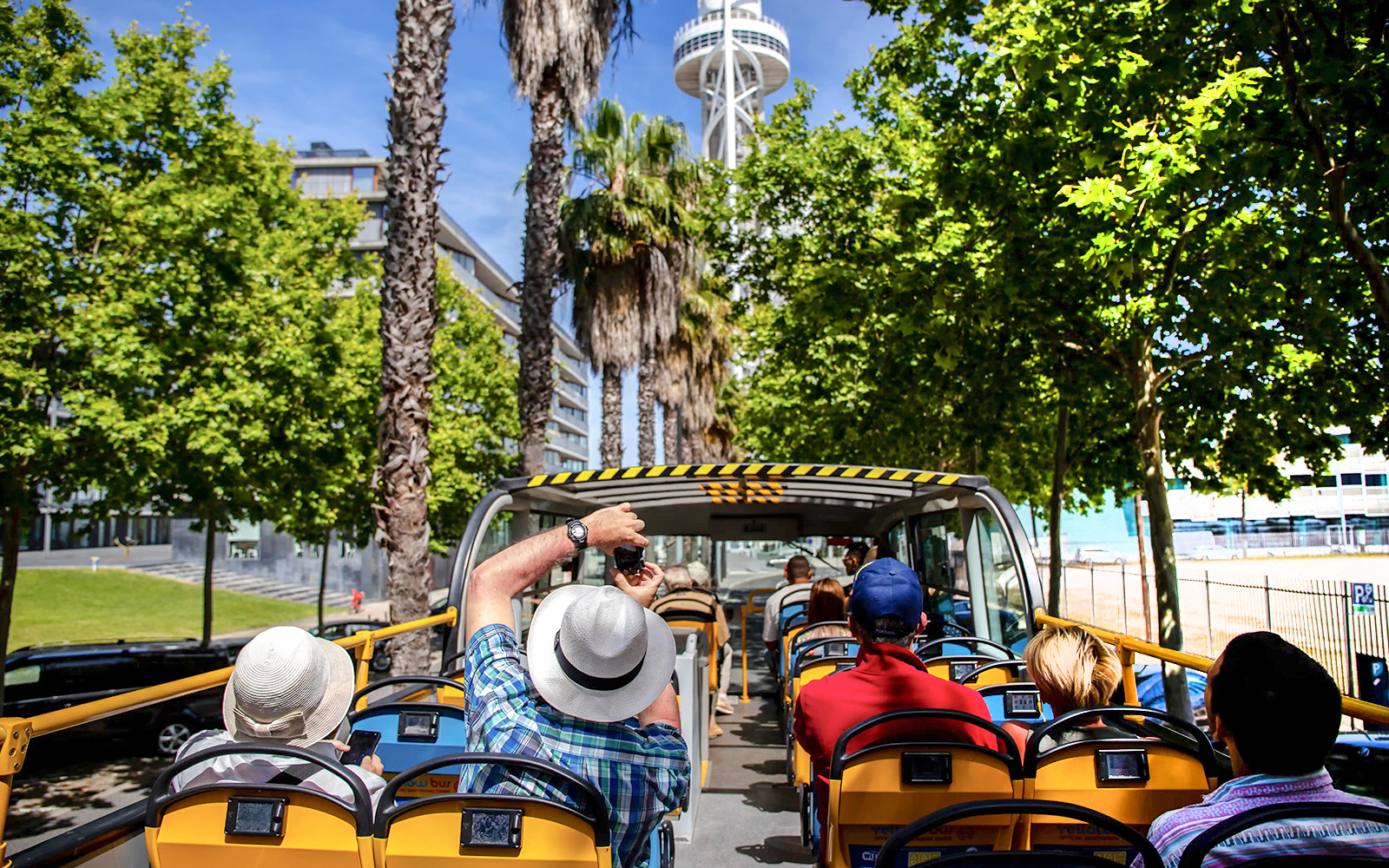 Tourists on an open-top yellow bus touring a city with a tall tower and palm trees.