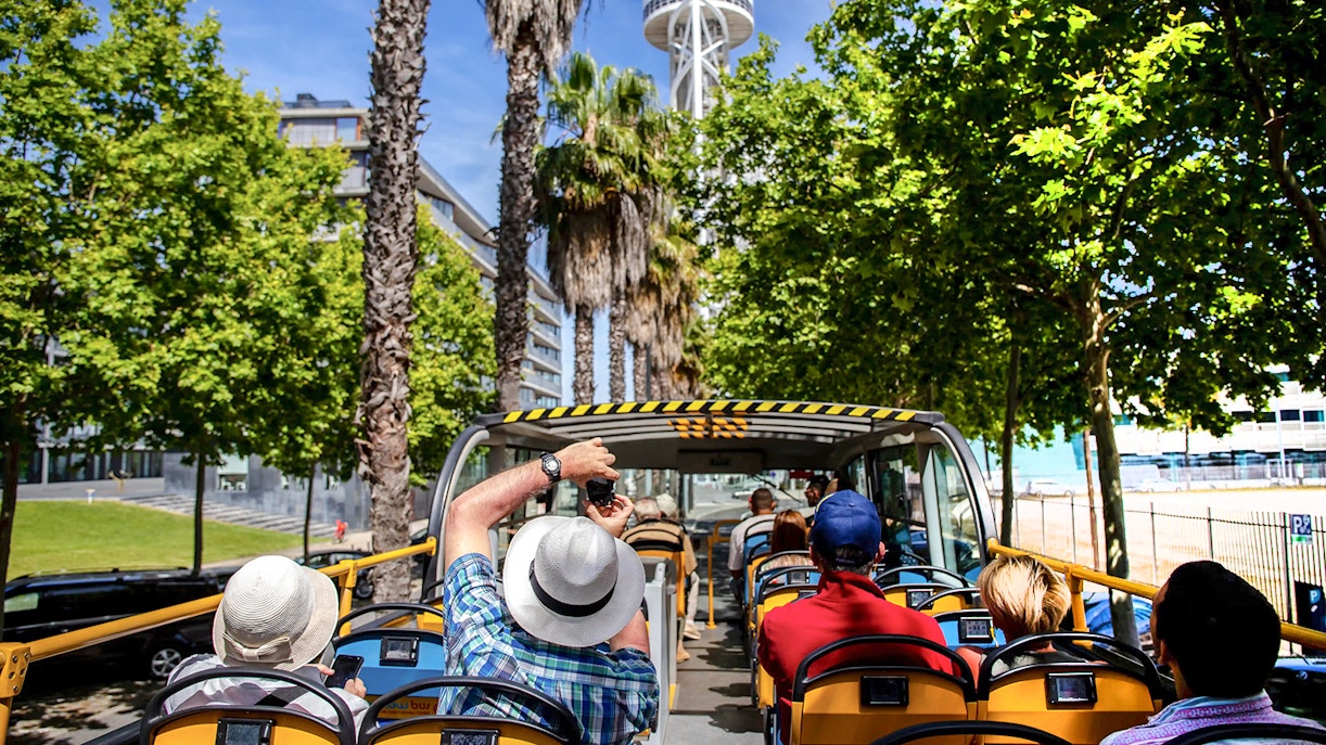 Tourists on an open-top yellow bus touring a city with a tall tower and palm trees.