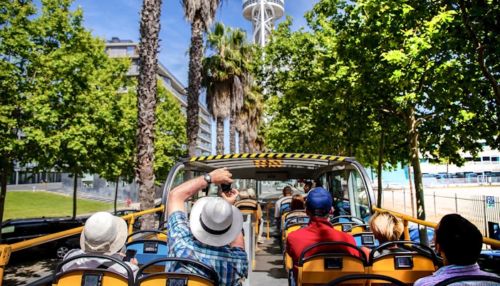 Tourists on an open-top yellow bus touring a city with a tall tower and palm trees.