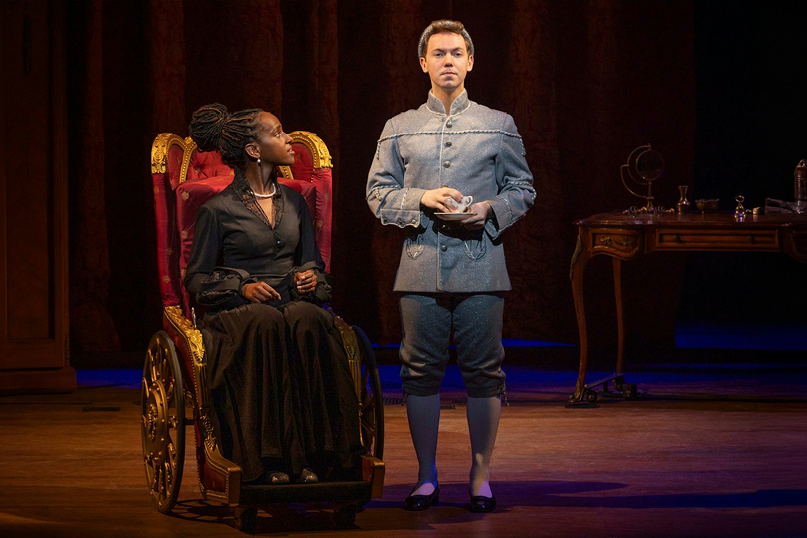 Stage scene from "Wicked" with two actors in period costumes, one seated, one standing.