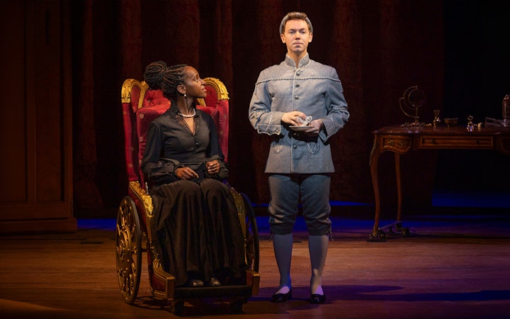 Stage scene from "Wicked" with two actors in period costumes, one seated, one standing.