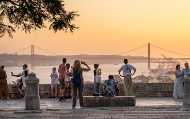 Visitors capturing sunset views from St George's Castle with Lisbon's bridge in the background.