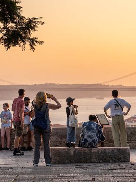 Visitors capturing sunset views from St George's Castle with Lisbon's bridge in the background.