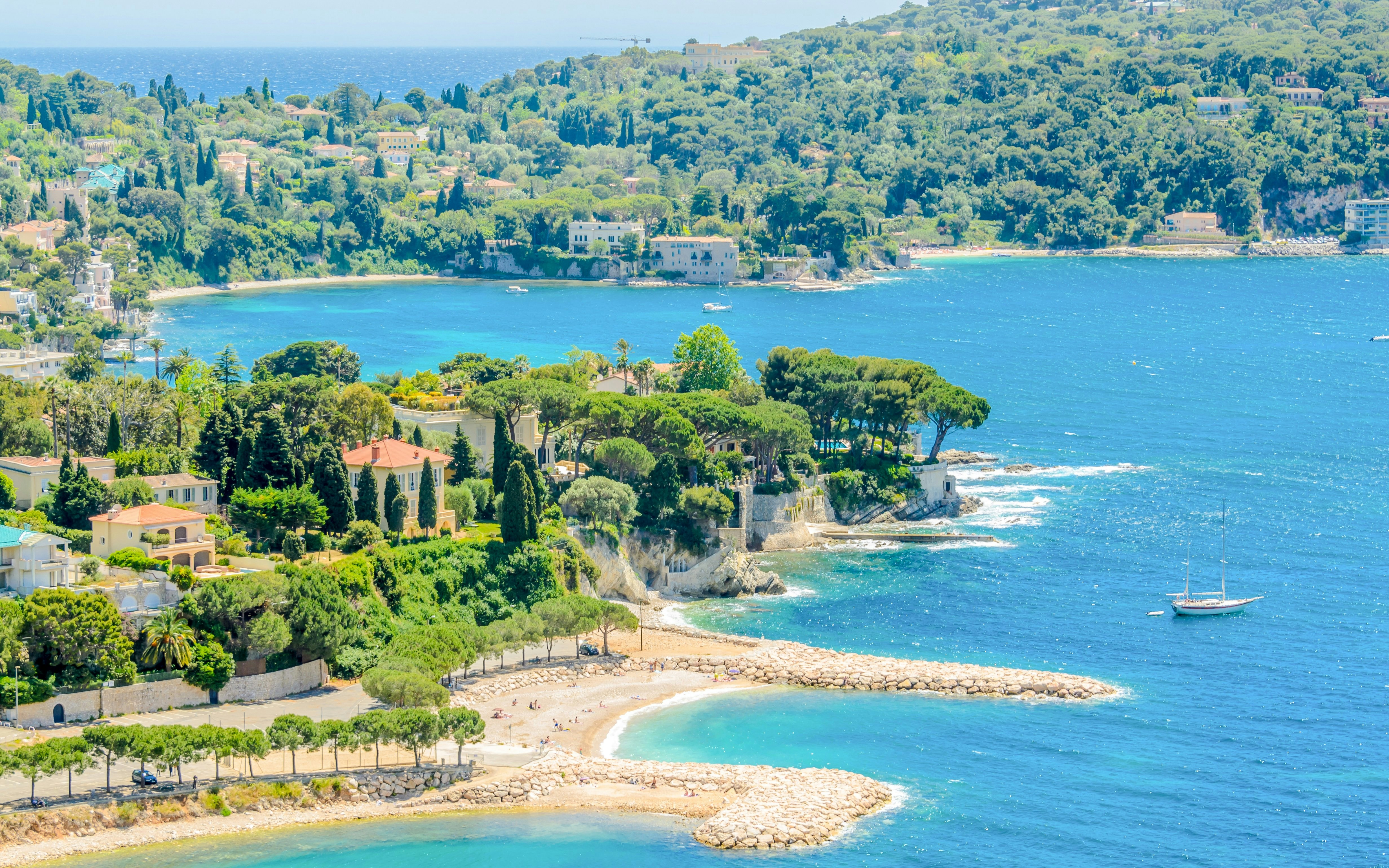 Coastal view of Villefranche-sur-Mer with lush greenery and a sailboat on the Mediterranean Sea.