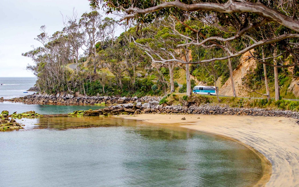 Stewart Island coastal road with tour bus near sandy bay and lush trees.