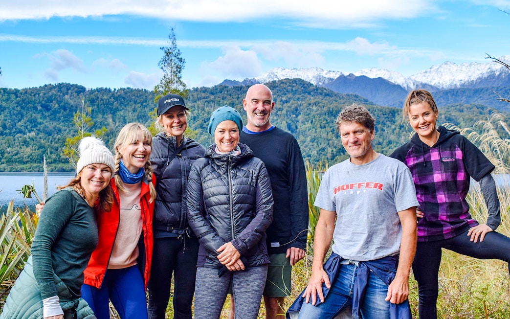 Group of tourists smiling during a rainforest walk at Franz Josef, with lush greenery and mountains in the background.