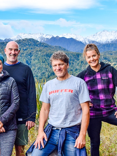 Group of tourists smiling during a rainforest walk at Franz Josef, with lush greenery and mountains in the background.