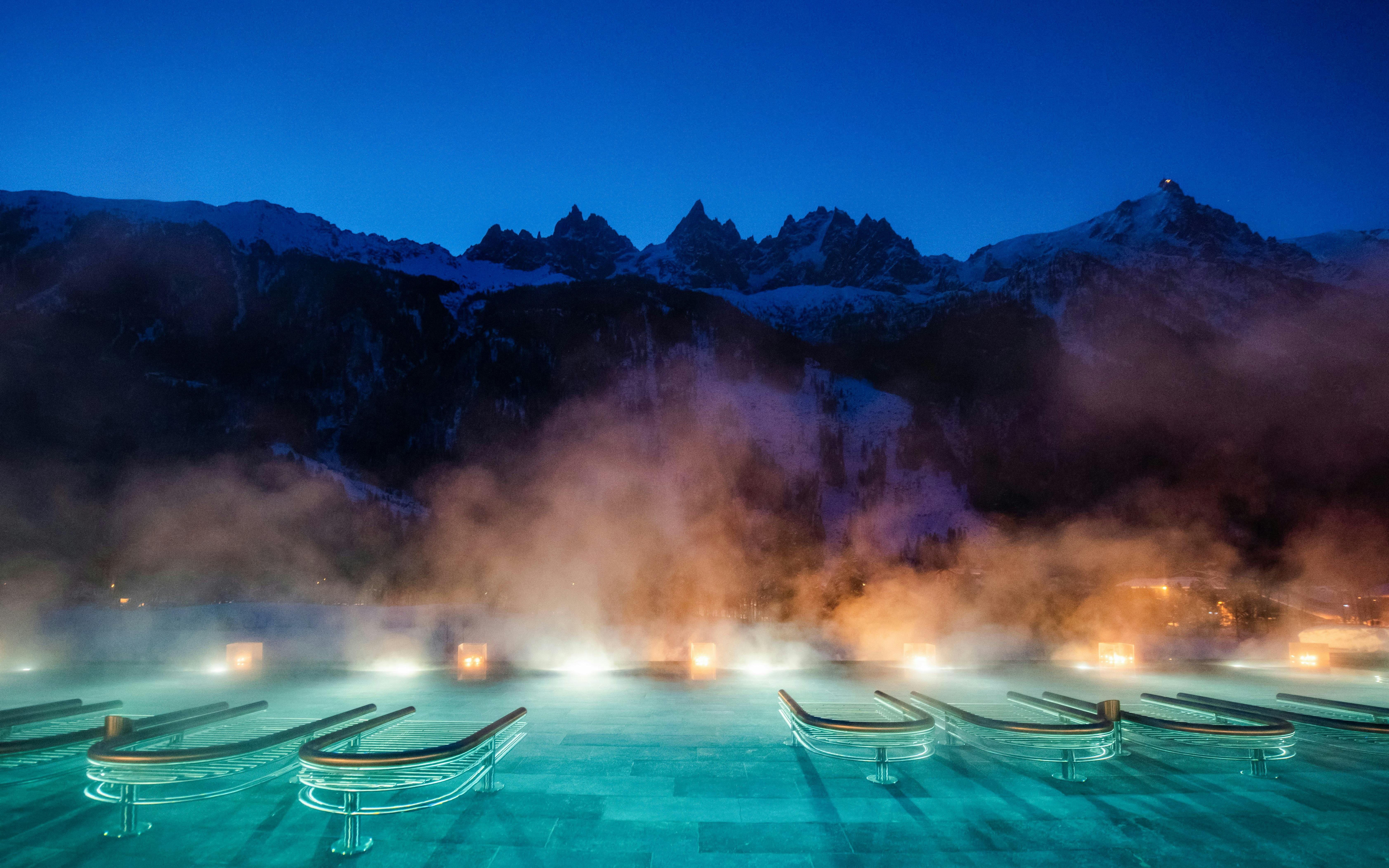 Outdoor thermal pool with steam at QC Terme Chamonix, framed by snow-capped mountains at dusk.