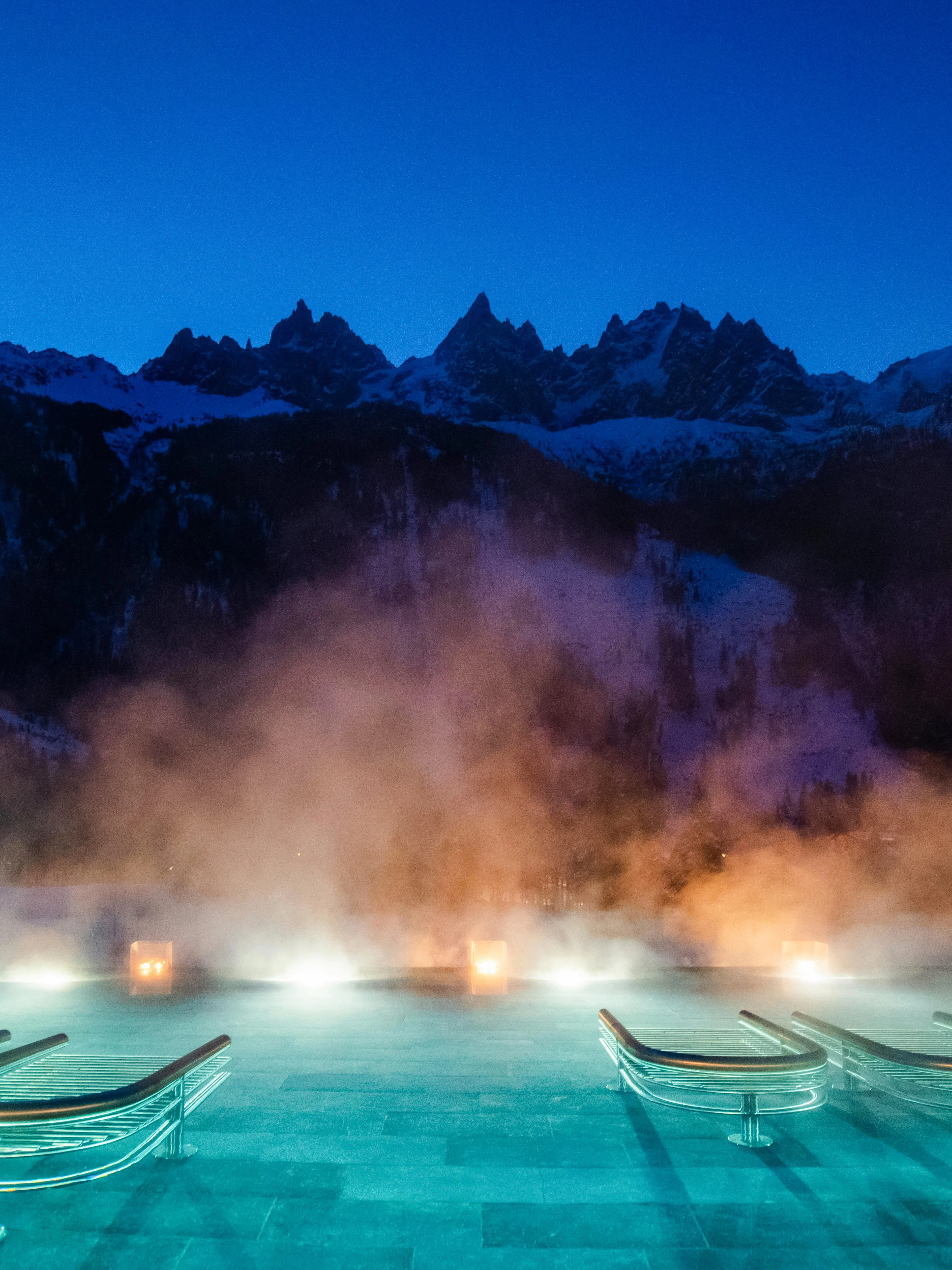 Outdoor thermal pool with steam at QC Terme Chamonix, framed by snow-capped mountains at dusk.
