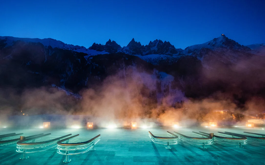 Outdoor thermal pool with steam at QC Terme Chamonix, framed by snow-capped mountains at dusk.