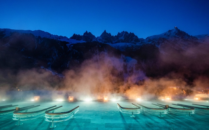Outdoor thermal pool with steam at QC Terme Chamonix, framed by snow-capped mountains at dusk.
