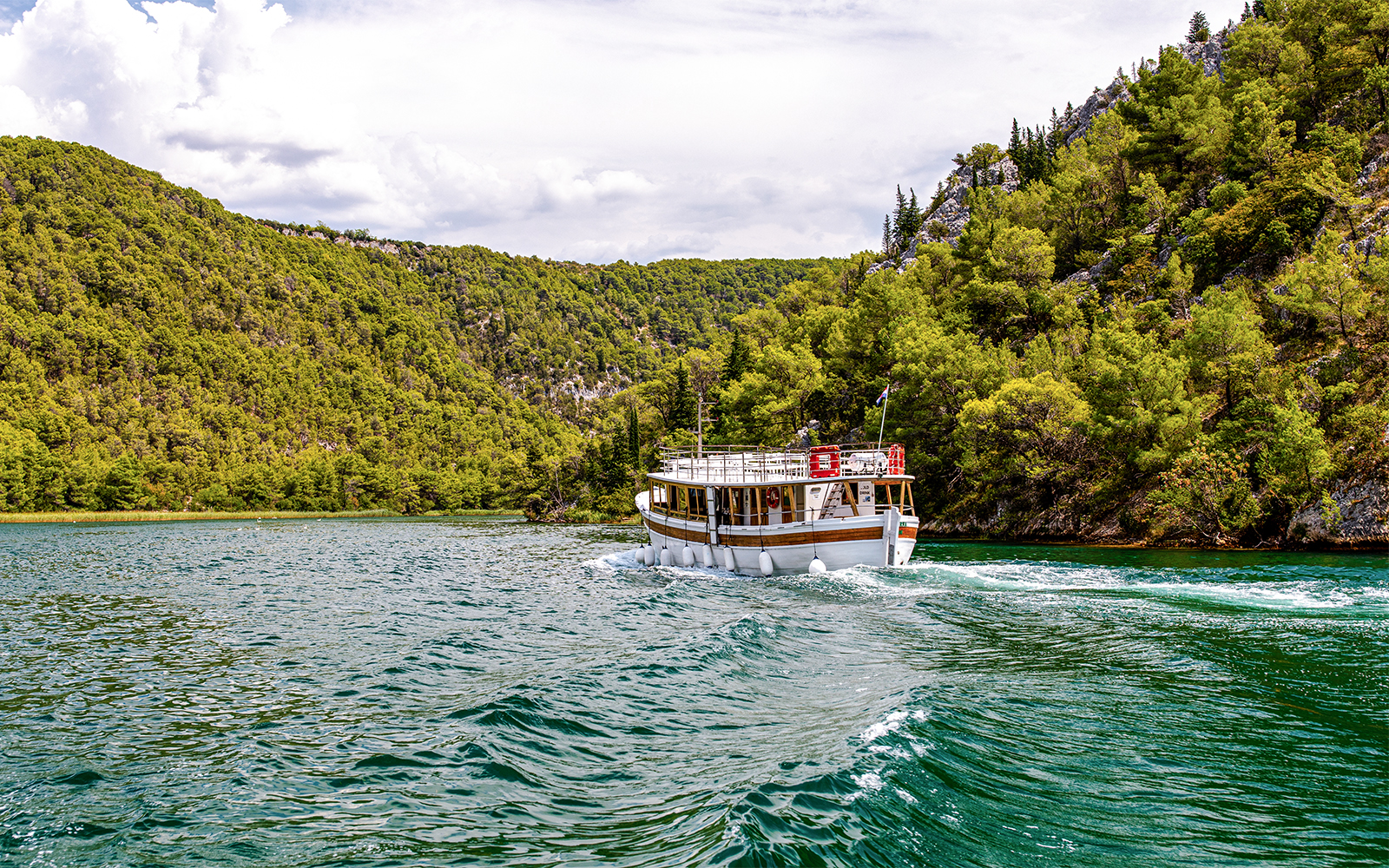 Boat cruising through Krka National Park surrounded by lush green hills.