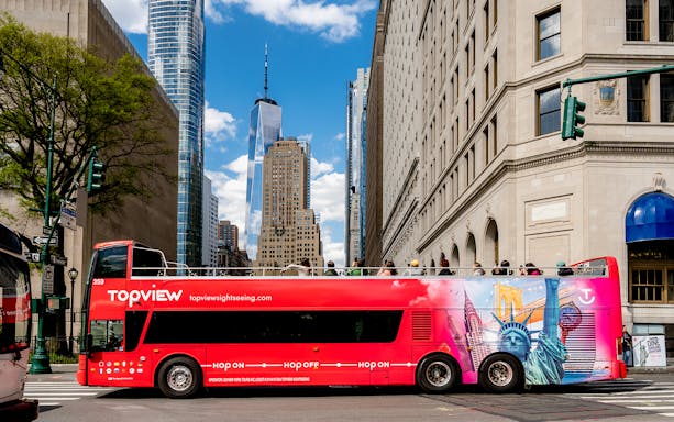 Red double-decker tour bus in New York City with One World Trade Center in background.