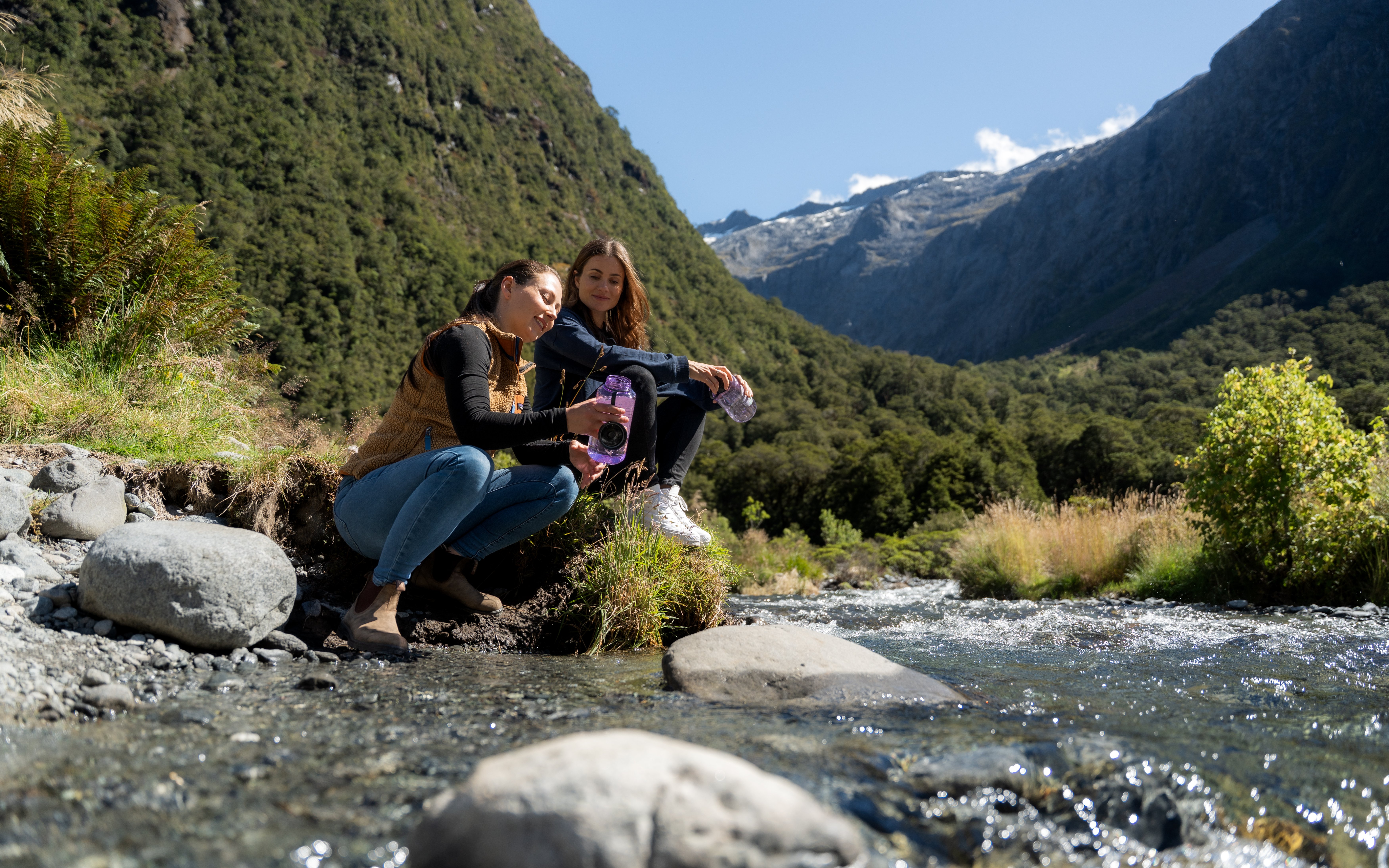 Two people sitting by a stream in Milford Sound with mountains in the background.
