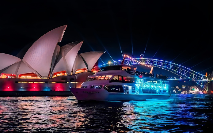 Cruise boat with neon lights near Sydney Opera House and Harbour Bridge at night.