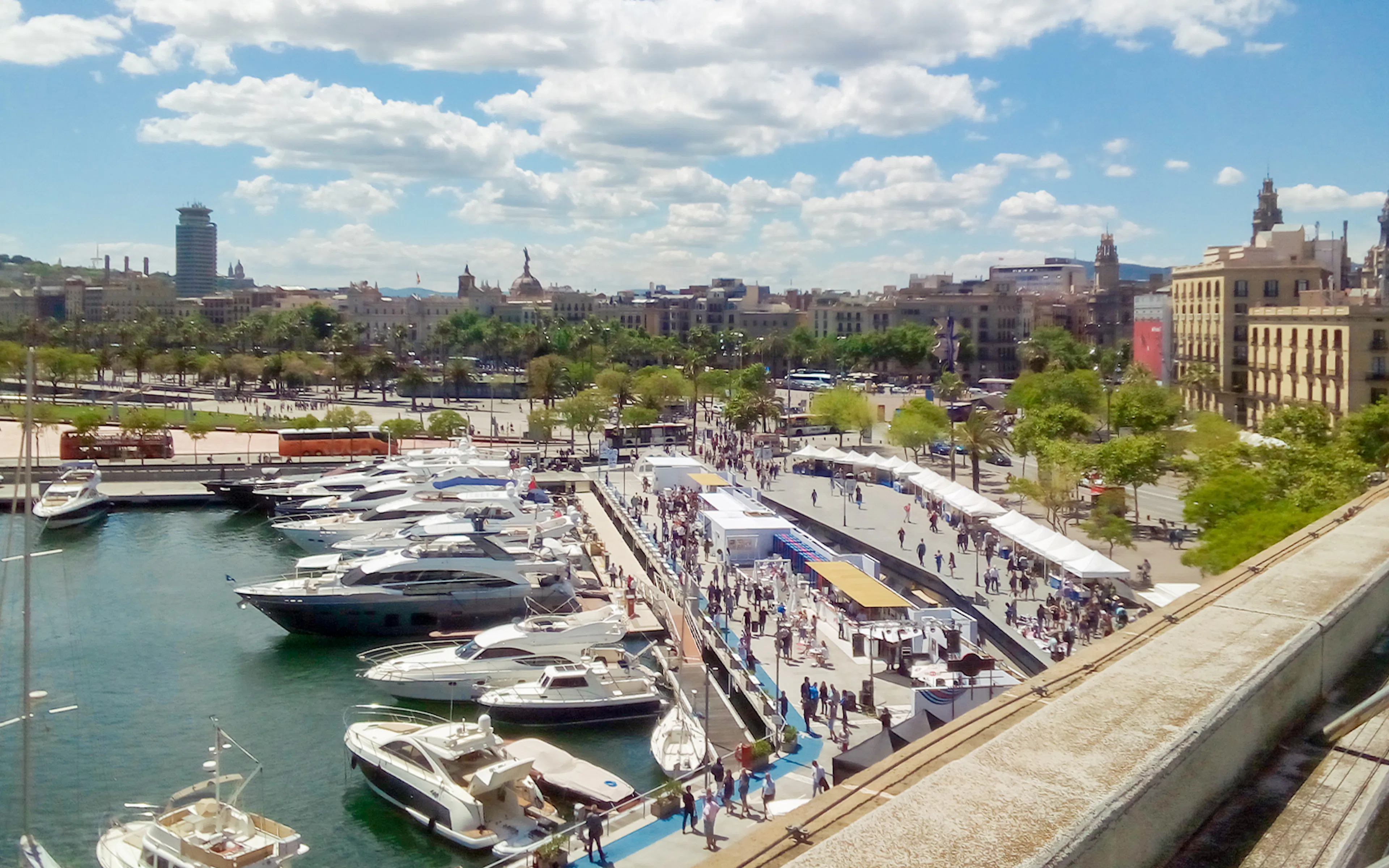 Yachts docked at Port Vell, Barcelona with cityscape and bustling promenade.