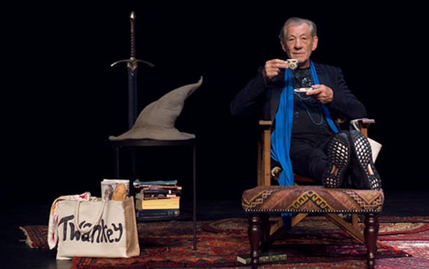 Actor on stage with a wizard hat, sword, and props, drinking tea.