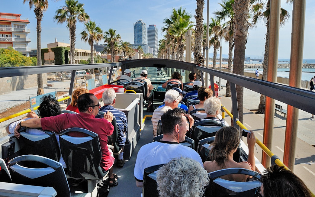 Tourists on an open-top bus enjoying a view of Barcelona's coastline and palm-lined streets.