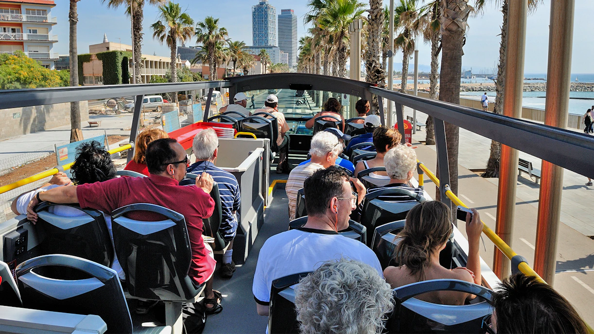Tourists on an open-top bus enjoying a view of Barcelona's coastline and palm-lined streets.