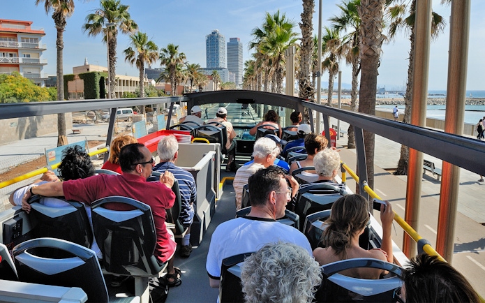 Tourists on an open-top bus enjoying a view of Barcelona's coastline and palm-lined streets.