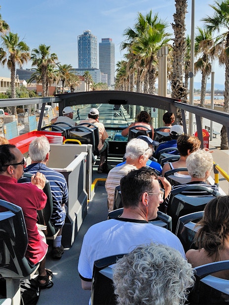 Tourists on an open-top bus enjoying a view of Barcelona's coastline and palm-lined streets.