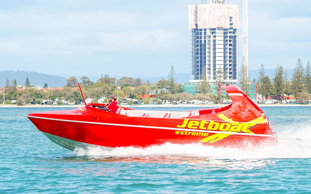 Red jet boat speeding on water near cityscape, part of Ultimate Jet Boat Ride and Surf Lesson.
