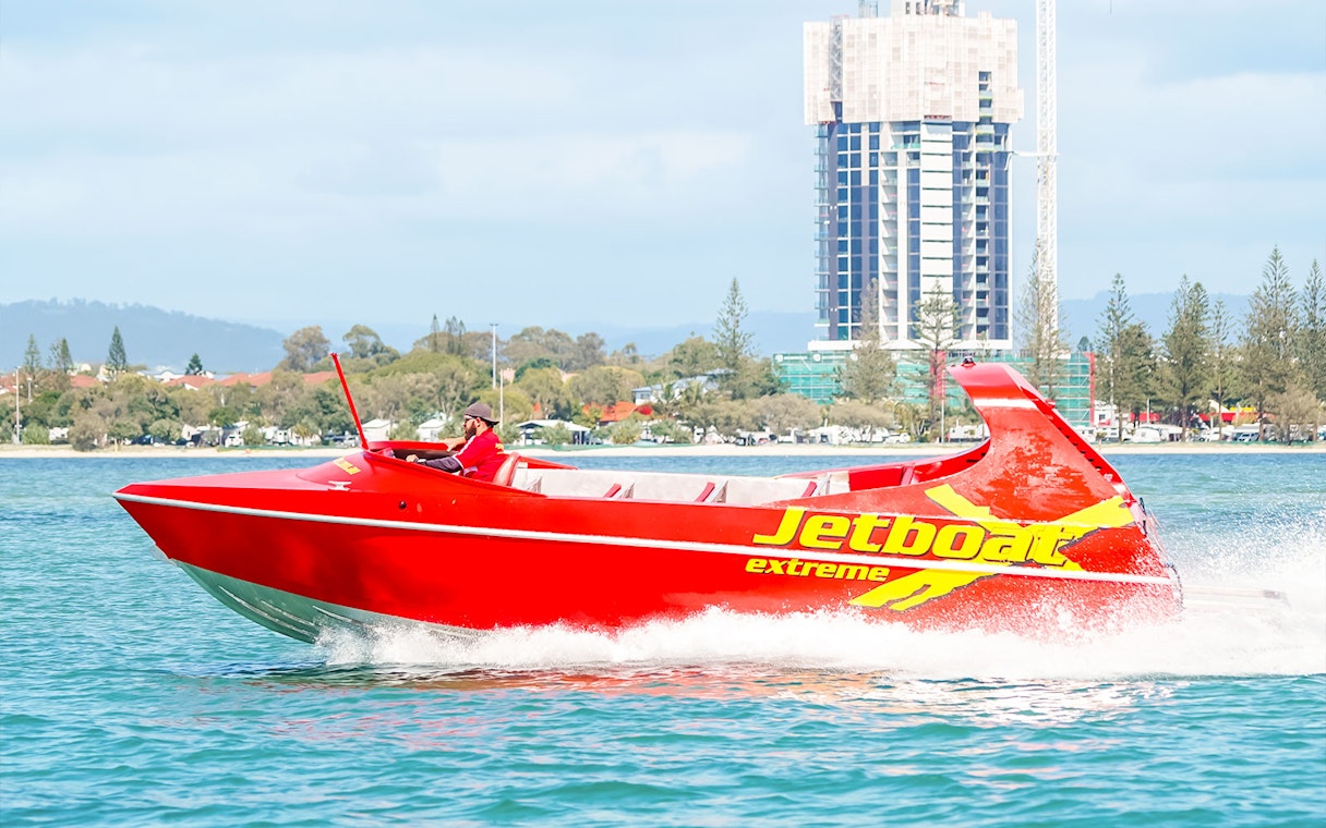 Red jet boat speeding on water near cityscape, part of Ultimate Jet Boat Ride and Surf Lesson.