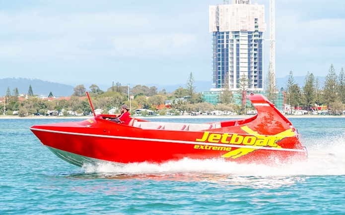 Red jet boat speeding on water near cityscape, part of Ultimate Jet Boat Ride and Surf Lesson.
