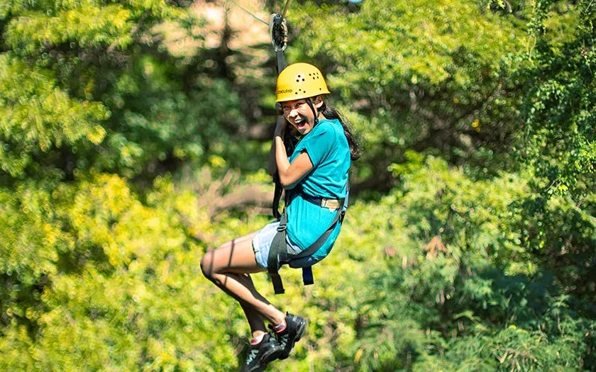 Person ziplining through forest with safety gear and helmet.