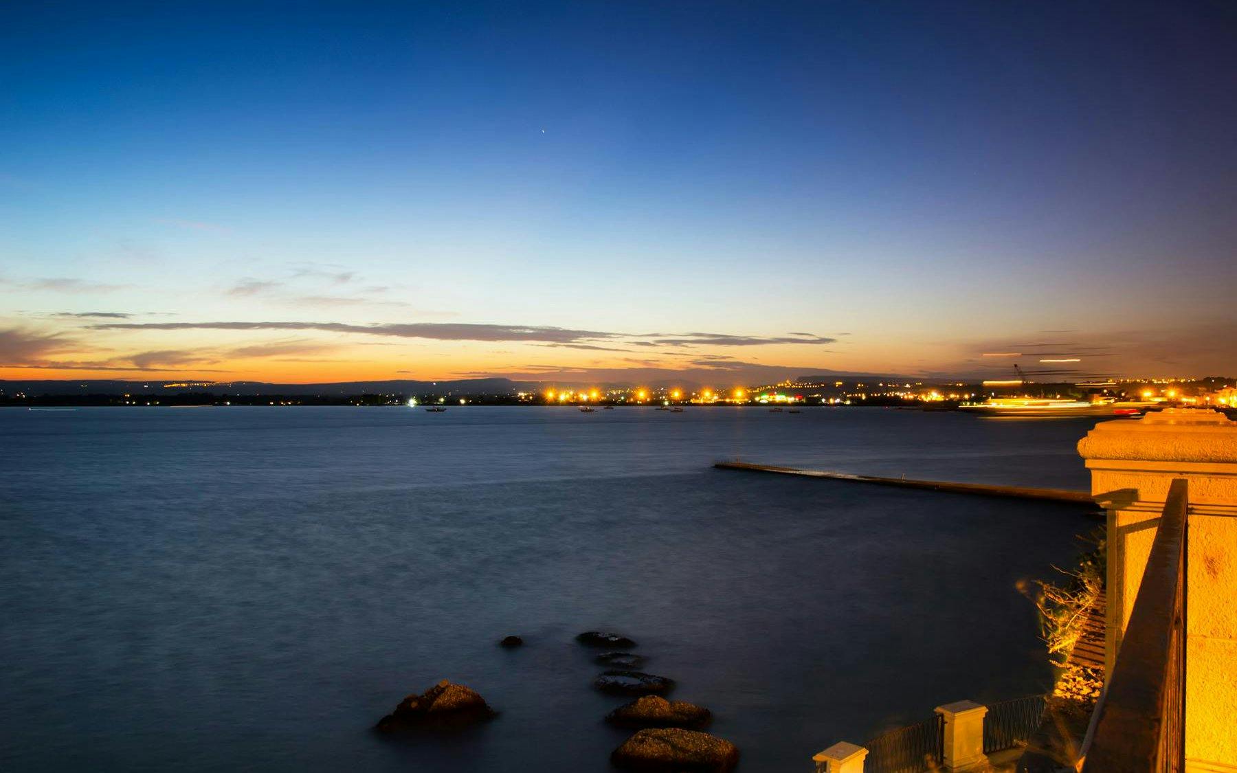 Ortigia sunset view over the sea with city lights in the distance.
