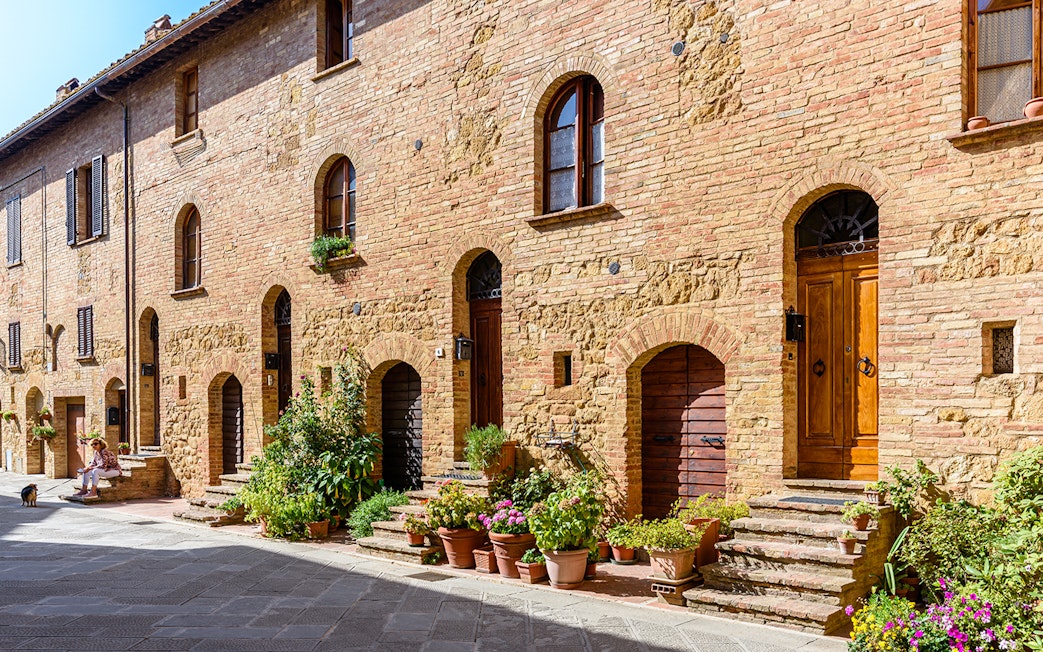 Historic brick buildings with arched doorways and potted plants in a sunlit Italian street.