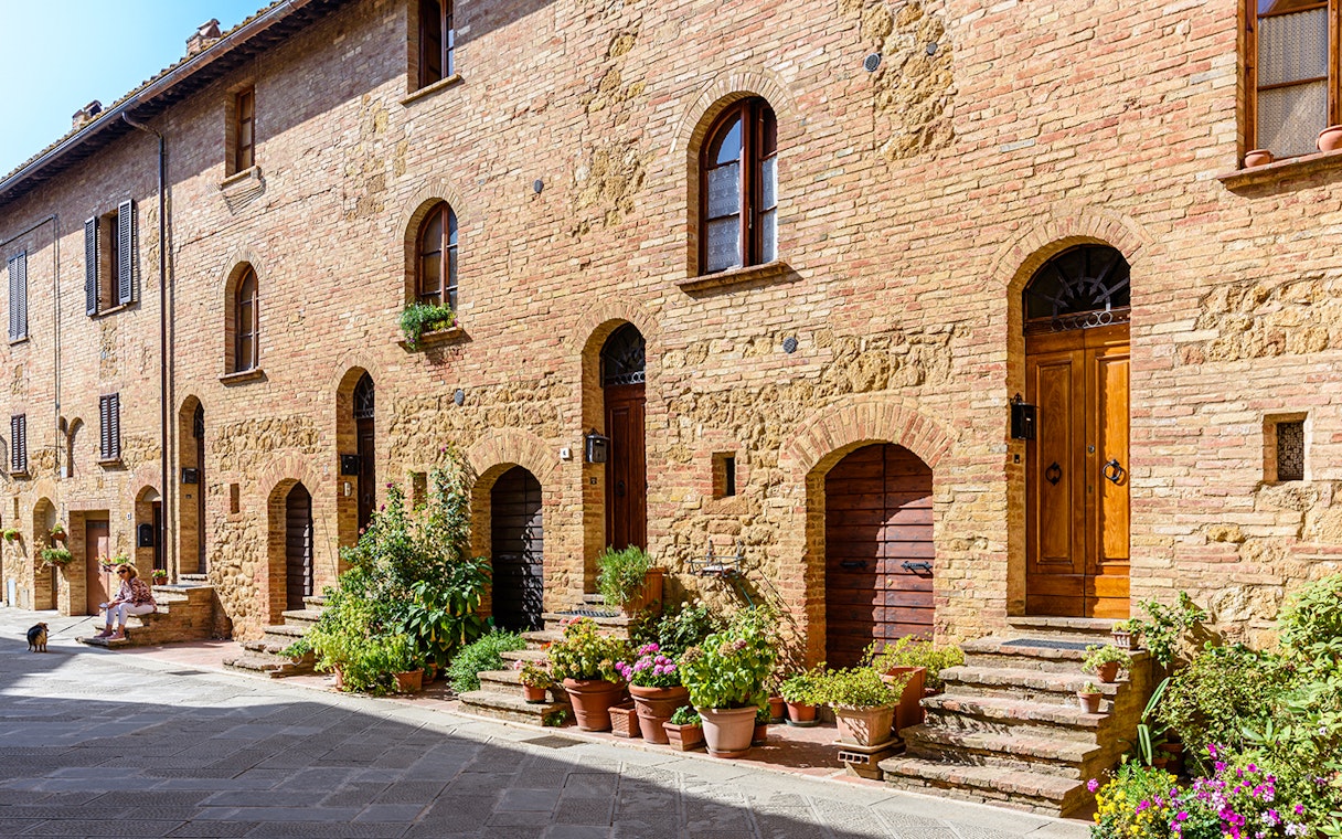 Historic brick buildings with arched doorways and potted plants in a sunlit Italian street.