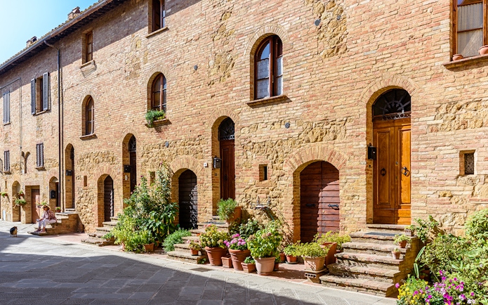 Historic brick buildings with arched doorways and potted plants in a sunlit Italian street.