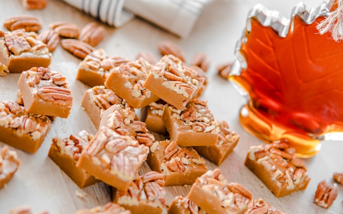 Maple fudge squares topped with pecans next to a maple syrup bottle.