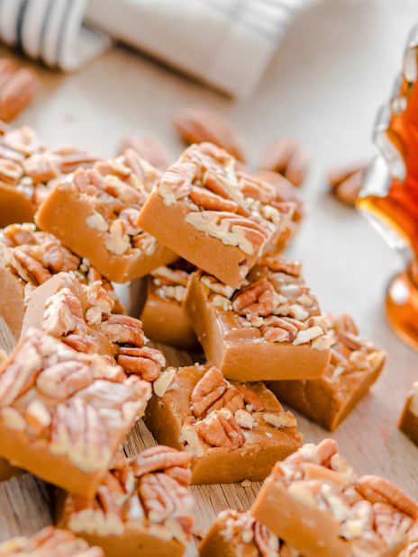 Maple fudge squares topped with pecans next to a maple syrup bottle.