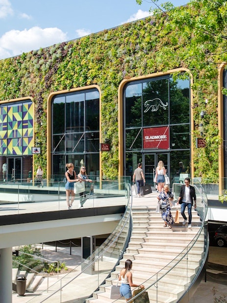 Shoppers walking at McArthurGlen Designer Outlet with green wall and colorful patterns.