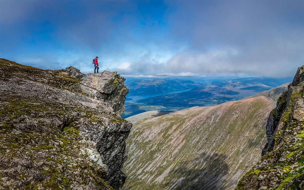 Hiker standing on rocky peak of Ben Nevis with expansive mountain views.