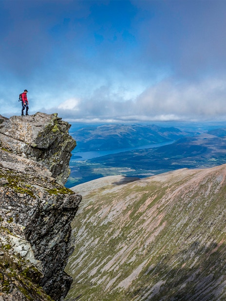 Hiker standing on rocky peak of Ben Nevis with expansive mountain views.