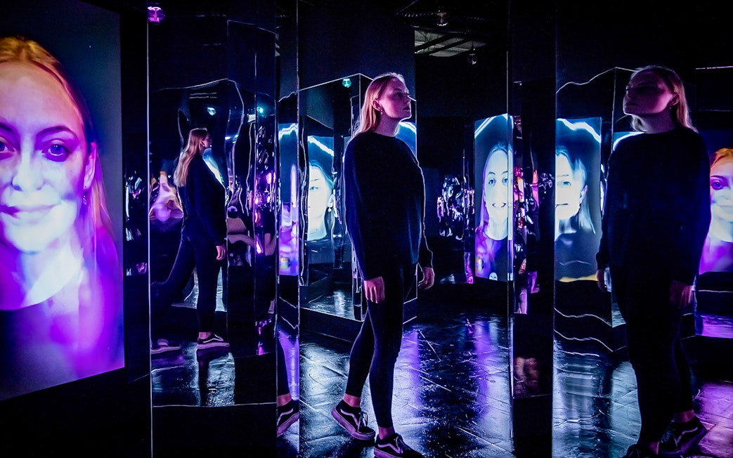 Person exploring mirrored room with digital faces at AMAZE Amsterdam Immersive Experience.