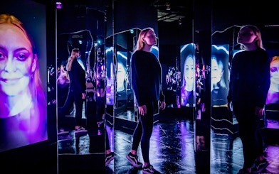 Person exploring mirrored room with digital faces at AMAZE Amsterdam Immersive Experience.