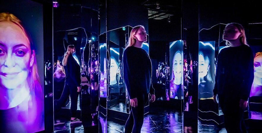 Person exploring mirrored room with digital faces at AMAZE Amsterdam Immersive Experience.