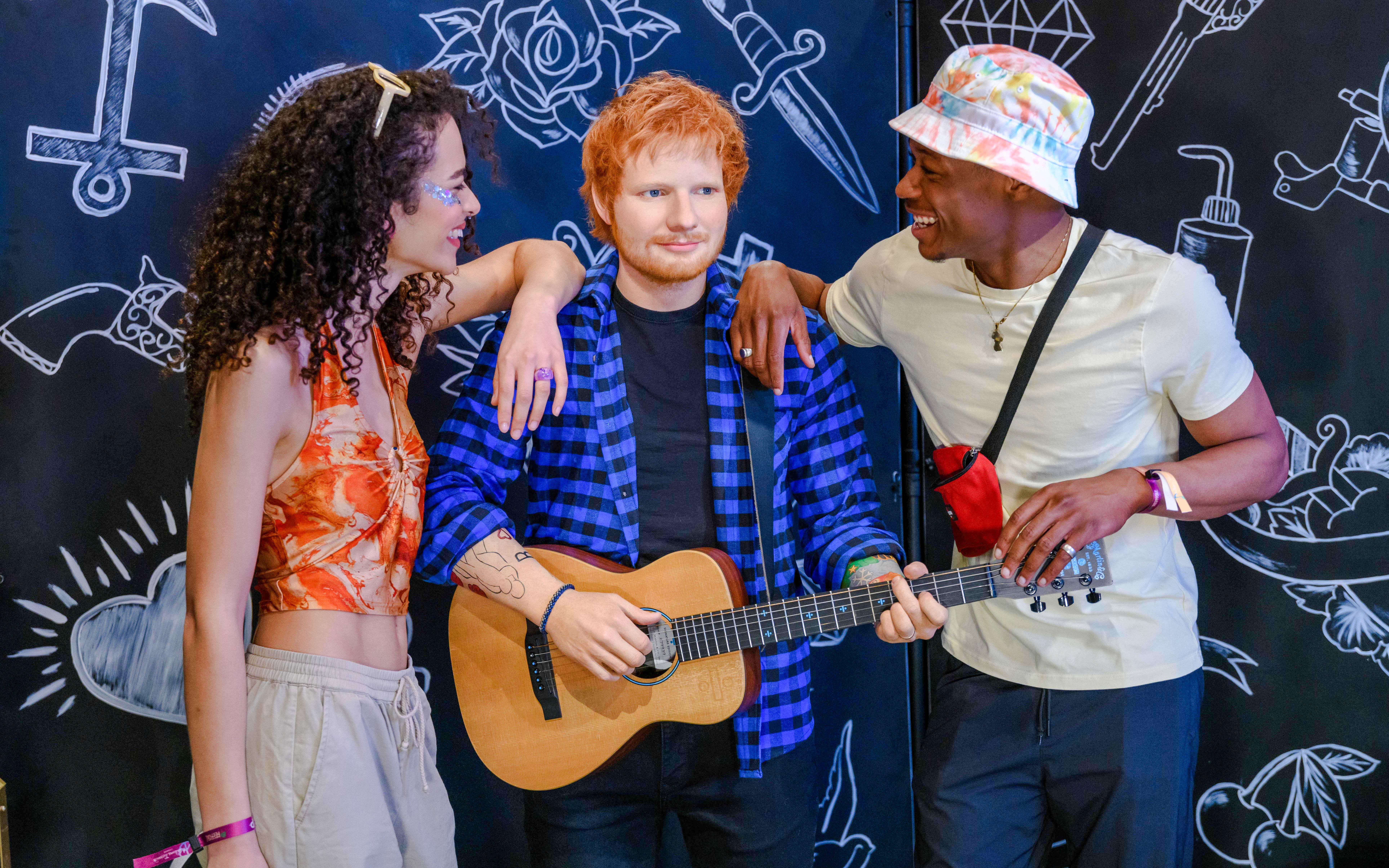 Wax figure of Ed Sheeran with guitar at Madame Tussauds London, flanked by two visitors.