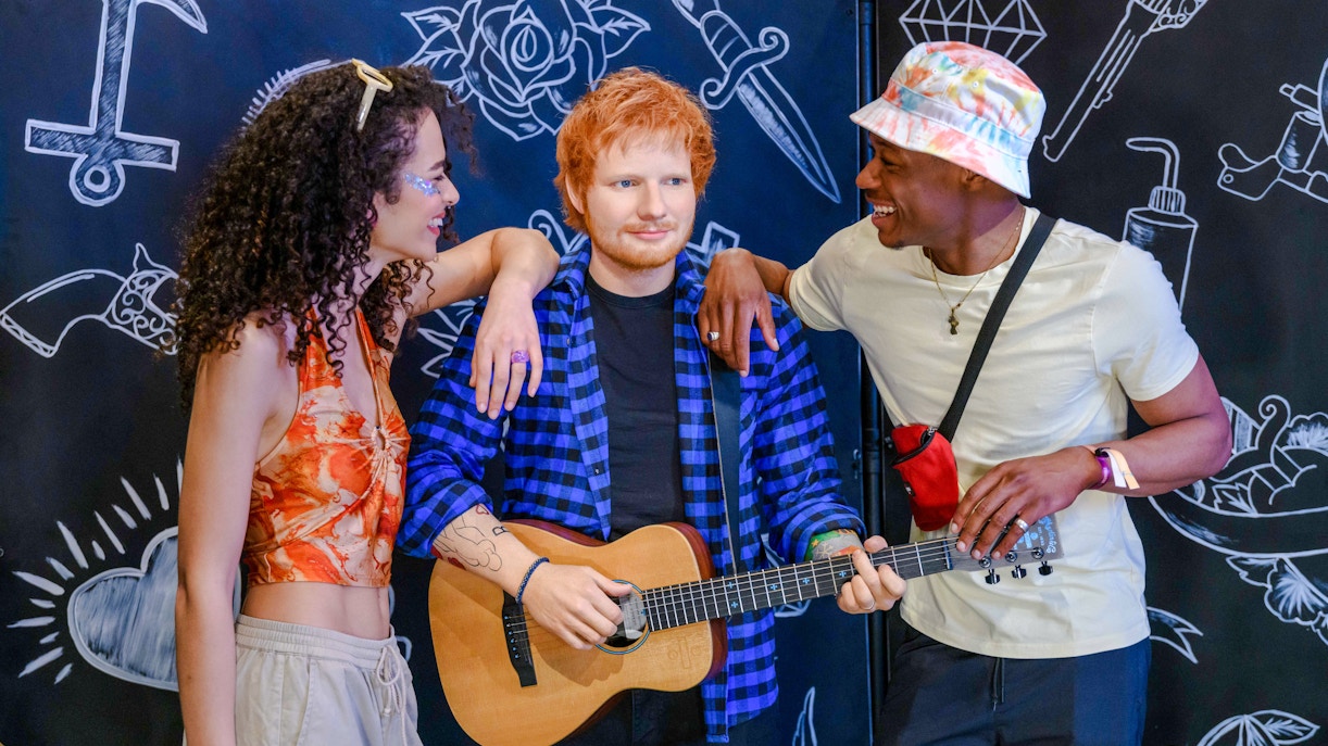 Wax figure of Ed Sheeran with guitar at Madame Tussauds London, flanked by two visitors.