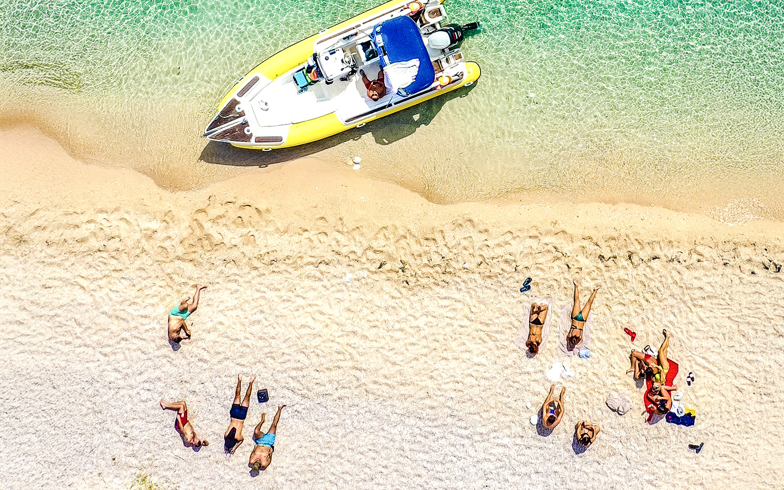 Aerial view of a small group relaxing on a sandy beach near Dafina Bay with a boat docked in clear water.