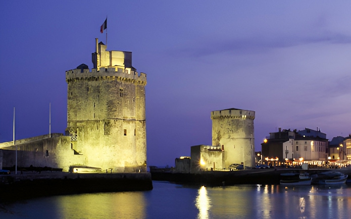 Towers of La Rochelle illuminated at dusk, reflecting on the water.
