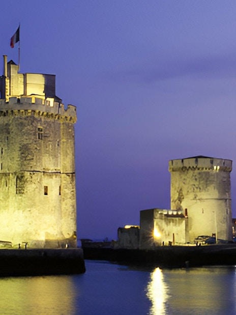 Towers of La Rochelle illuminated at dusk, reflecting on the water.